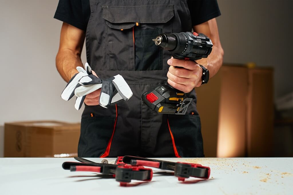 Radnik drži aku bušilicu i zaštitne rukavice iznad radnog stola