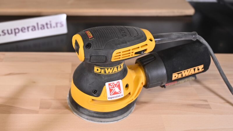 DeWalt ekscentar brusilica stoji na drvenoj površini sa priključenom vrećicom za prašinu